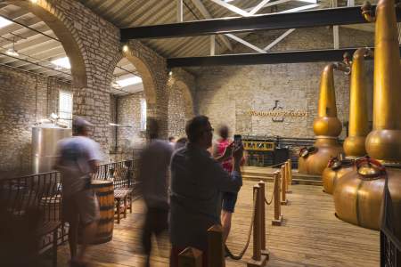 A candid photo of people taking a tour of one of the rooms of Woodford Reserve’s Distillery Tour. It’s a tall room with white brick walls, and three large bronze-colored metal devices used in the bourbon production process. The Woodford Reserve name and logo can be seen straight ahead on the wall.